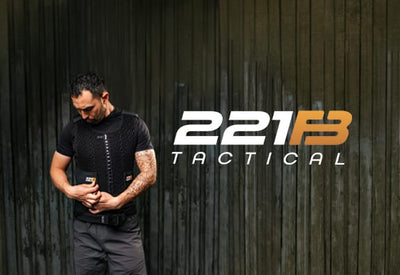 A man with a beard and tattoos wears a black tactical vest, adjusting it against a dark wood wall. The white and orange 