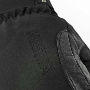 Black mitten by Hestra with an embossed HESTRA logo on textile fabric and a textured leather panel with stitching.