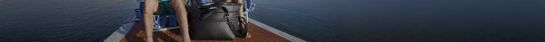 Legs of two people and a black AO brand bag on a sunny wooden boat deck, overlooking dark blue water.