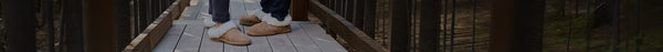 Two pairs of feet in tan and white fluffy slippers stand on a light-colored wooden deck, flanked by dark railings and a blurry forest.