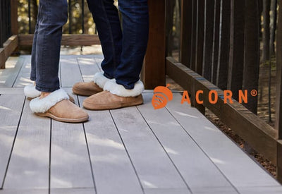 Two people in dark jeans and tan fur-lined slippers stand comfortably on a light wooden deck. An orange ACORN® logo is in the bottom right.