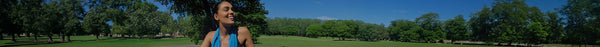 A happy woman in a blue top smiles on the left, looking into a sunny, green park with numerous trees under a clear blue sky.