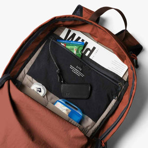 A lightweight rust-colored daypack by Bellroy, its interior showing an organized compartment with a magazine, snack, black pouch, white container, and blue kit.