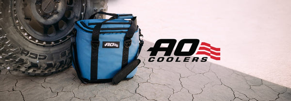 A blue AO Coolers soft cooler bag rests on cracked dry ground beside a large off-road tire. The AO Coolers logo is on the right.