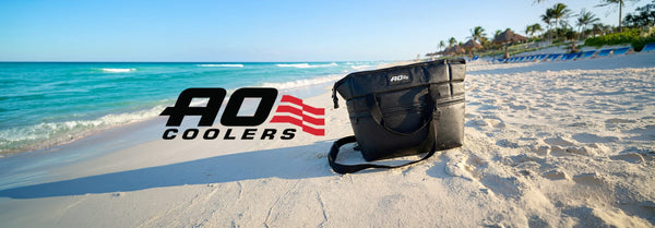 A black AO Coolers bag rests on a sunny white sand beach beside turquoise ocean waves. 