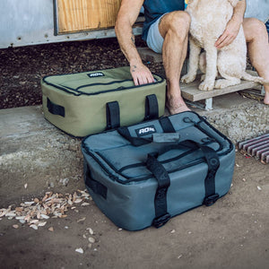 A man with a dog on his lap sits by two soft coolers, one green and one grey. Both coolers feature an "AO" logo; the grey one's straps say "AO & GO".