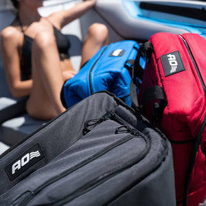 Three soft-sided cooler bags, gray, blue, and red, are in the foreground, each with a white "AO=" logo. A woman in a black bikini relaxes on a boat in the blurred background.