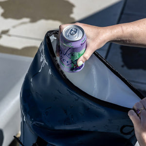 A hand with light green nail polish places a purple La Croix Blackberry Cucumber sparkling water can into a dark blue, wet cooler bag.