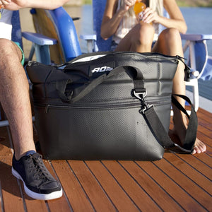 A black carbon fiber cooler bag with an "AO" logo sits on a wooden deck between a man's leg and a woman relaxing with a drink.