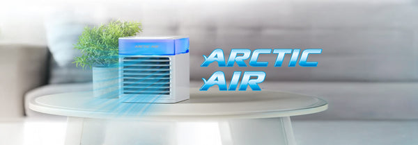 A white Arctic Air personal cooler with blue light emits cool air next to a potted plant on a light table, with the 