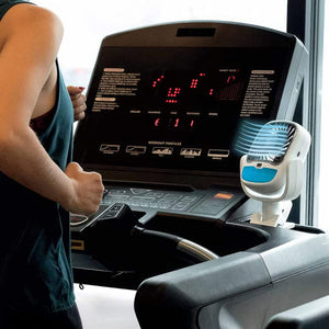 A person in a tank top runs on a black treadmill console displaying red digital metrics and workout profiles. A small white and blue fan is clipped to the right handlebar.