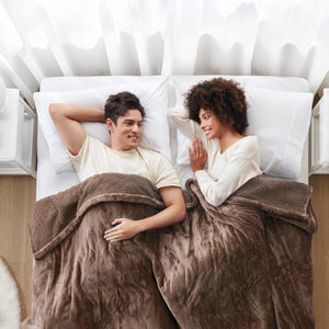 A diverse couple smiles at each other while lying in bed under separate fluffy brown blankets.