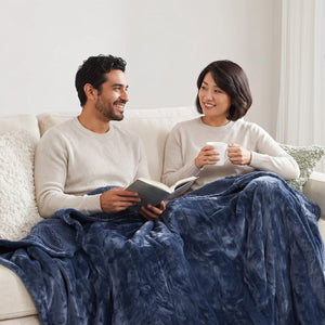 A smiling man reads a book beside a smiling woman holding a mug, both sharing a blue blanket on a cream sofa.