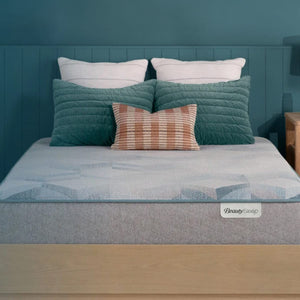 A well-made bed with a gray BeautySleep mattress, teal headboard, white pillows, a teal quilted pillow, and a striped accent pillow.