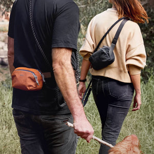 From behind, a man with an orange fanny pack and a woman with a black sling bag walk outdoors, both handling dark rope leashes, with a dog partially visible.