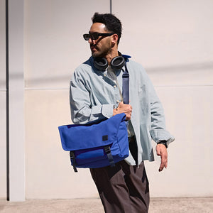 A man in sunglasses and a light blue shirt walks right, wearing headphones and holding a royal blue messenger bag in a sunny outdoor setting.