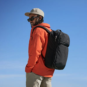A smiling man in an orange shirt, light cap, and sunglasses looks left, wearing a black Bellroy backpack against a bright blue sky.