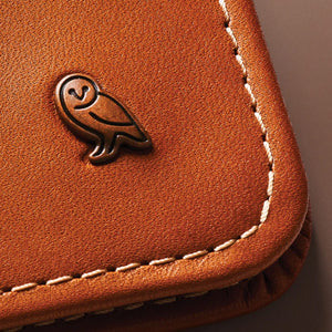 Close-up of textured brown leather with a bronze owl logo and visible white stitching along the curved edge.