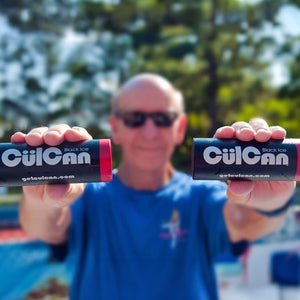 A smiling man in sunglasses and a blue shirt holds two black CülCan Black Ice cylindrical products, each with a red band and "getculcan.com" visible, in an outdoor setting.