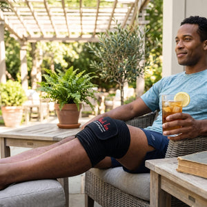 A Black man relaxes on a sunny patio, wearing a Black Ice knee brace on his left knee and holding iced tea. Potted plants and a pergola are in the background.