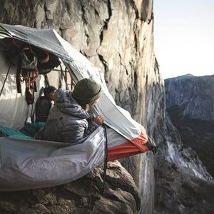 A person in a beanie and puffer jacket looks out of a portaledge tent anchored to a sheer rock cliff, revealing a vast mountain landscape.