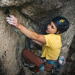 A focused woman in a yellow BLACK shirt and Black Diamond helmet places a cam while rock climbing on a textured rock face.