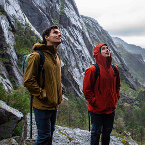 Two hikers in brown and red jackets with backpacks stand on rocks, looking up at towering grey mountains under an overcast sky.