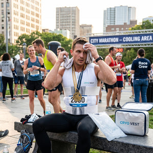 A male marathon runner with a medal and a white CRYOVEST sits on a bench, wiping his face. A CRYOVEST cooler bag is nearby, with a "CITY MARATHON - COOLING AREA" sign behind him.