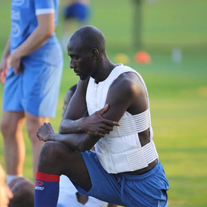 A dark-skinned man in a white padded vest and blue shorts kneels on a grassy field, looking down. Other blurred figures and green grass are in the background.