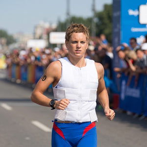 A female athlete with short blonde hair runs on a sunny road, wearing a white padded vest, blue/red shorts, and a "10" on her left arm. Spectators line the blurred background.