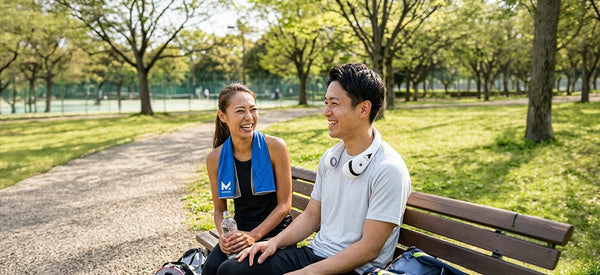 Two cheerful Asian adults sit on a park bench. The woman has a blue 'M' logo towel, the man a white neck fan, with a sunny park and tennis courts behind them.