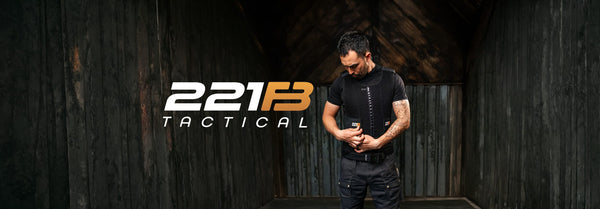 A man in a black mesh tactical vest adjusts it in a dark wooden room, with the white and orange 