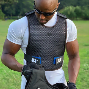 A bald man in sunglasses wears a white shirt and black mesh vest with white "XX" logos, looking down while holding black gear outdoors.