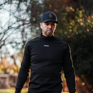 A man in a black turtleneck and baseball cap with "221B" logo looks down, bathed in warm sunlight against a blurred outdoor background.