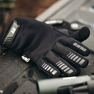 A black left glove lies on a dark textured surface, showing a grey "F" logo on the cuff, reflective silver stripes on fingers, and "221B" text on the index fingertip.