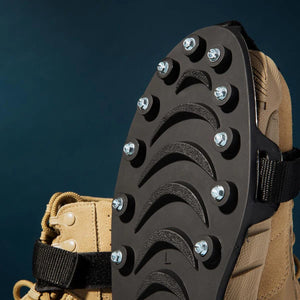Close-up of a black ice gripper sole with metal studs and textured treads strapped to a tan hiking boot, against a dark blue background.