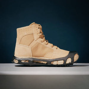 A tan tactical boot, featuring a black spiked ice grip wrapped around its sole, sits on a white surface against a dark blue background.