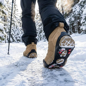 A person in black pants and tan boots walks on a snowy path; the right boot shows a black ice gripper with red studs. Sunny winter forest.