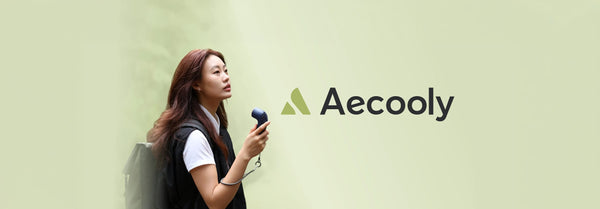 A young woman in a white tee and black vest holds a dark portable fan, looking up against a light green wall, with the 