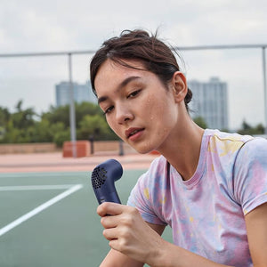 A sweaty young woman with damp hair wears a tie-dye shirt and holds a dark blue handheld fan on an outdoor sports court.