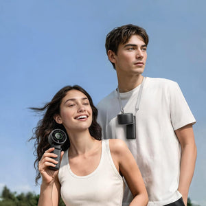 A smiling woman holds a handheld fan showing '100' while a man wears a neck fan, both outdoors under a clear blue sky.