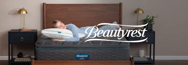 A woman sleeps comfortably on a dark gray Beautyrest mattress with teal accents in a stylish bedroom. The Beautyrest logo is prominently displayed.
