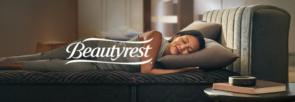 A woman sleeps peacefully on her side on a dark patterned Beautyrest mattress, illuminated by sunlight. A grey upholstered headboard is on the right.