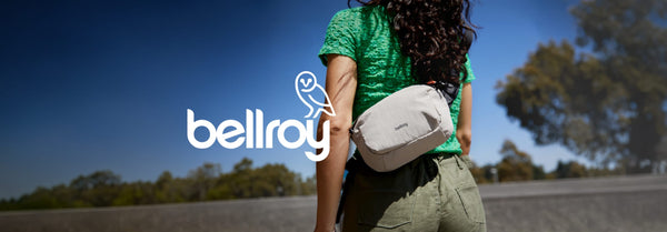 A person from behind, wearing a green top and olive pants, carries a light grey Bellroy sling bag. A large white Bellroy logo with an owl is on the left, against a blue sky.