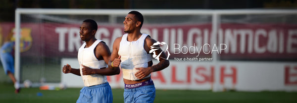 Two smiling male athletes in white monitoring vests jog on a soccer field with a blurred red banner and the 