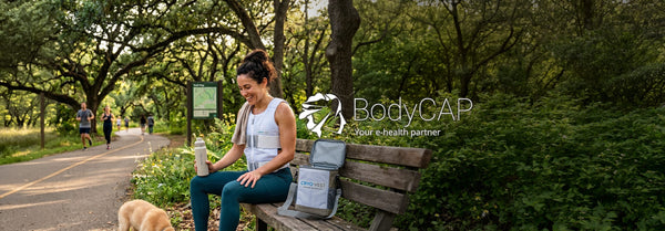 Smiling woman in a cooling vest on a park bench with a Cryo Vest cooler, holding a bottle. Joggers are on a tree-lined path behind her. BodyCAP logo overlaid.
