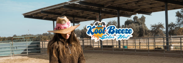 A woman in a straw hat with a fan and pink scarf stands in a sunny horse arena, with a 