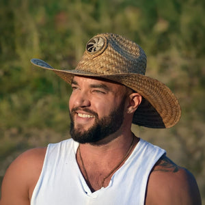 A smiling man with a dark beard, wearing a white tank top and a straw cowboy hat with a front-mounted fan, looks left against a blurry green background.