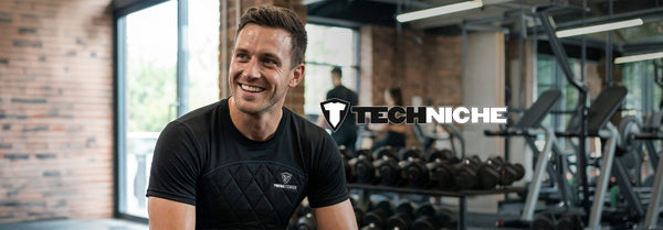 A smiling, sweaty man in a black quilted Techniche t-shirt at a gym. Racks of dumbbells and equipment are blurred in the background, with a large 