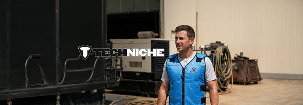 A man in a blue cooling vest stands outdoors in a sunny industrial area with machinery and a beige building, featuring a 